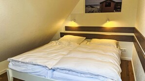 Un lit simple avec une literie blanche, dans une pièce dont le plafond est en pente et où l’on aperçoit une petite image d’un abri de plage accrochée au mur.