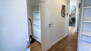Un couloir aux murs blancs, doté d’un plancher en bois et d’une porte blanche avec un trou de serrure. On y trouve un porte-manteau avec divers articles et une petite étagère sur laquelle sont posés des papiers.