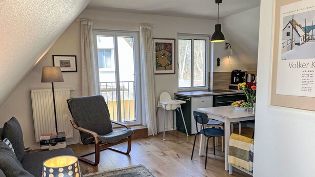Un salon confortable avec un canapé, un fauteuil, une table à manger avec des chaises, un petit coin cuisine et une grande fenêtre avec des rideaux.