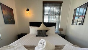 A neatly made bed with white linens and a rolled towel, a wall-mounted light, and a window with blinds.