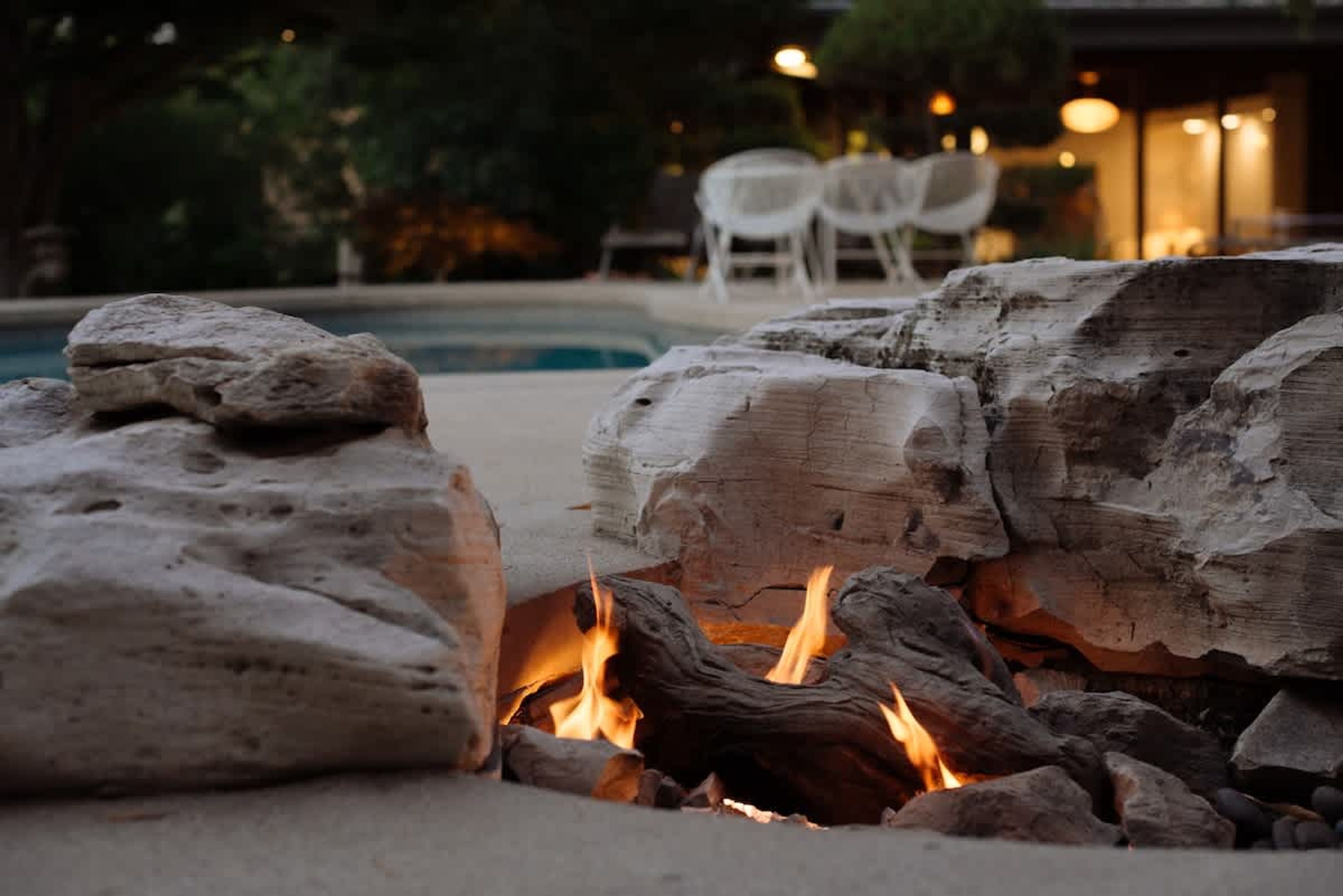 A stone fire pit with logs and flames, surrounded by a poolside area with chairs and a building in the background.