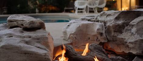 Un pozo de fuego de piedra con leña y llamas, rodeado de una zona de piscina con sillas y un edificio al fondo.