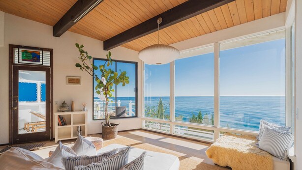 A modern living room with a large window overlooking the sea, a cozy sofa, and a wooden ceiling.