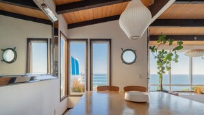 A dining area with a wooden table, chairs, and a large window offering a view of the ocean.