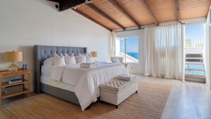 A bedroom with a large bed, wooden headboard, and a view of the sea through a window.
