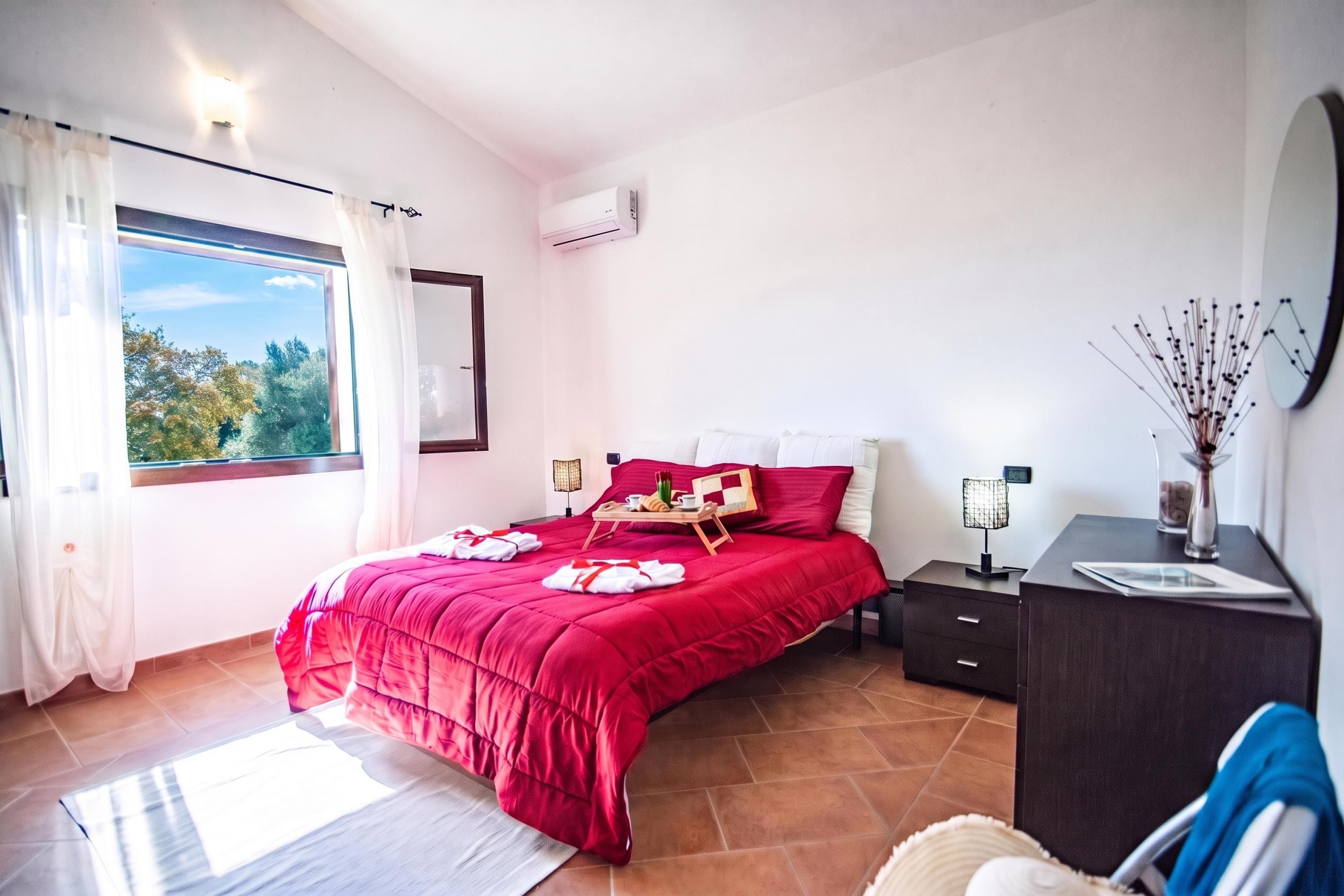Une chambre à coucher avec un grand lit, une table de chevet, une commode, une fenêtre avec des rideaux et une vue sur les arbres à l’extérieur.