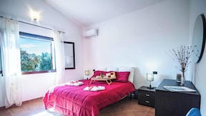 Un dormitorio con un ventanal grande, una cama con colcha roja, una mesita de noche con lámpara y un aparador.