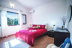 Quarto com cama grande, janela com cortinas, mesa de cabeceira com abajur e cômoda.