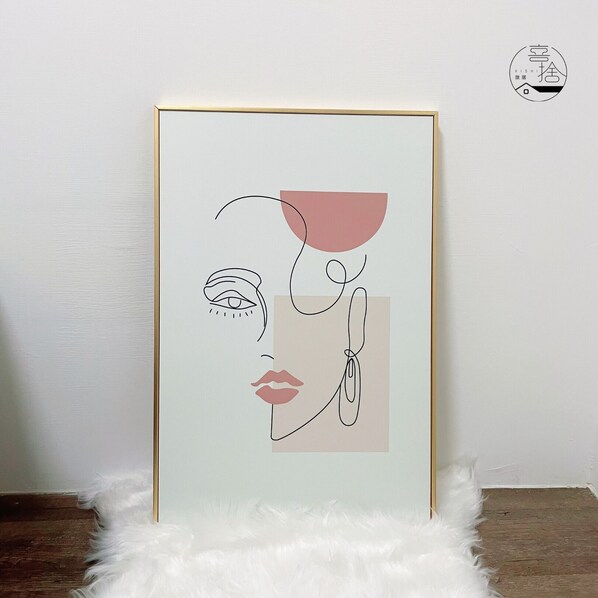 Une œuvre d’art encadrée, représentant des formes humaines abstraites, est exposée sur un tapis blanc et moelleux, devant un mur uni.