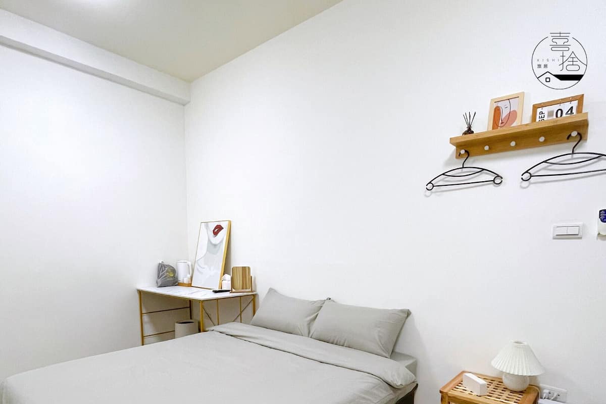 A bedroom with a bed, a desk, a lamp, and a small wooden shelf with items on it.
