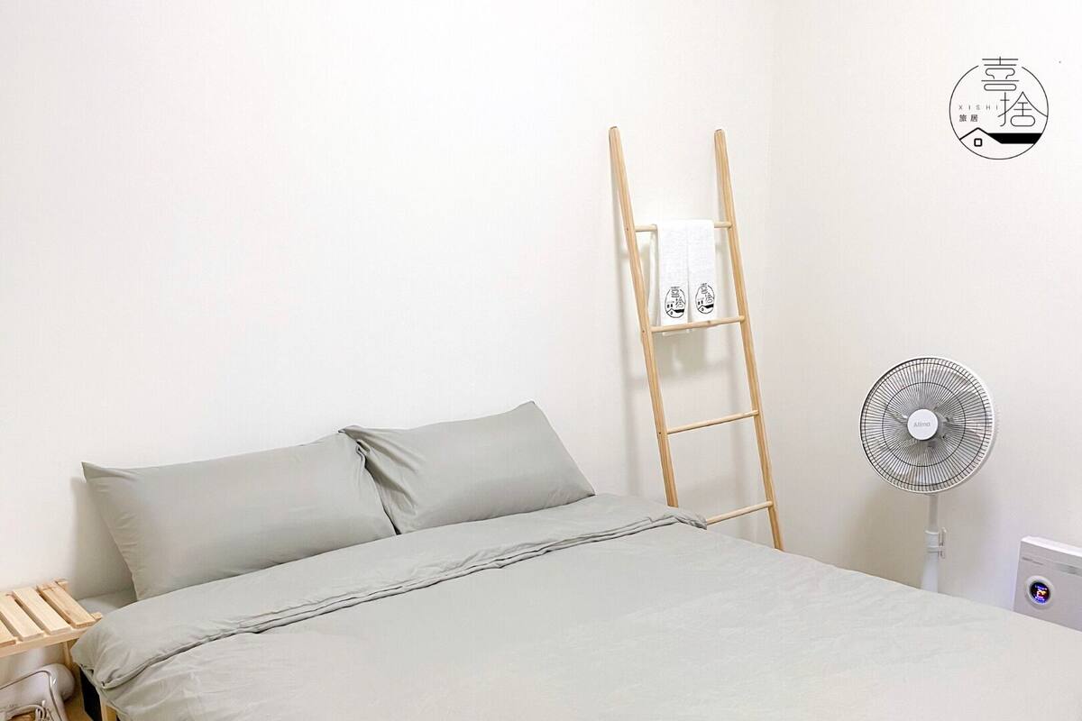 A bed with a grey mattress and pillows, a wooden ladder leaning against the wall, and a standing fan on the floor.