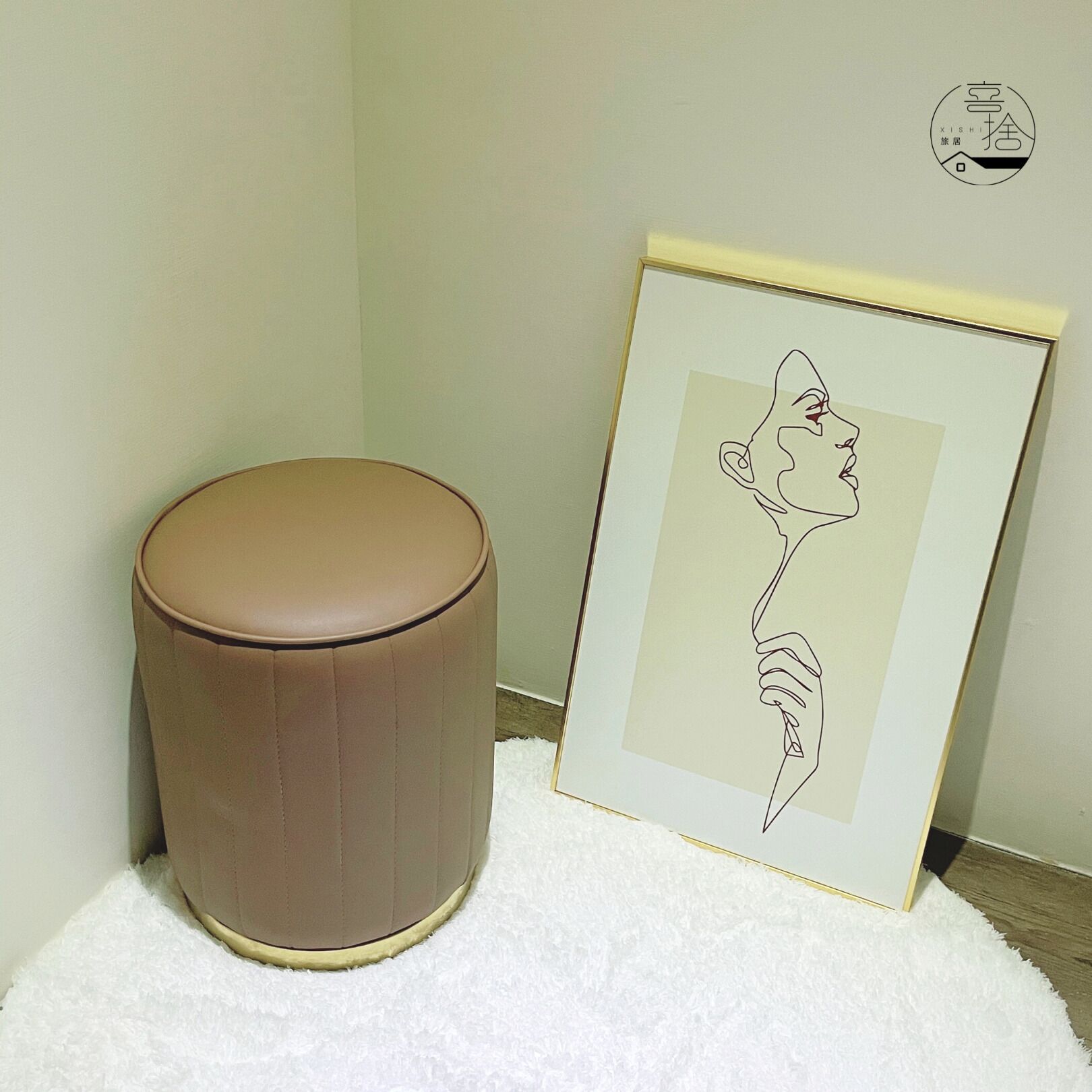 A framed artwork of a stylized animal head and a brown cylindrical stool on a white textured rug.