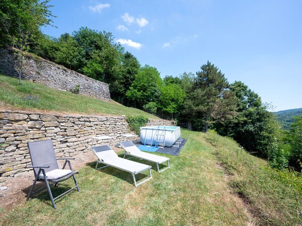 A stone wall, two lounge chairs, and a hot tub in a grassy outdoor area with trees and hills in the background.