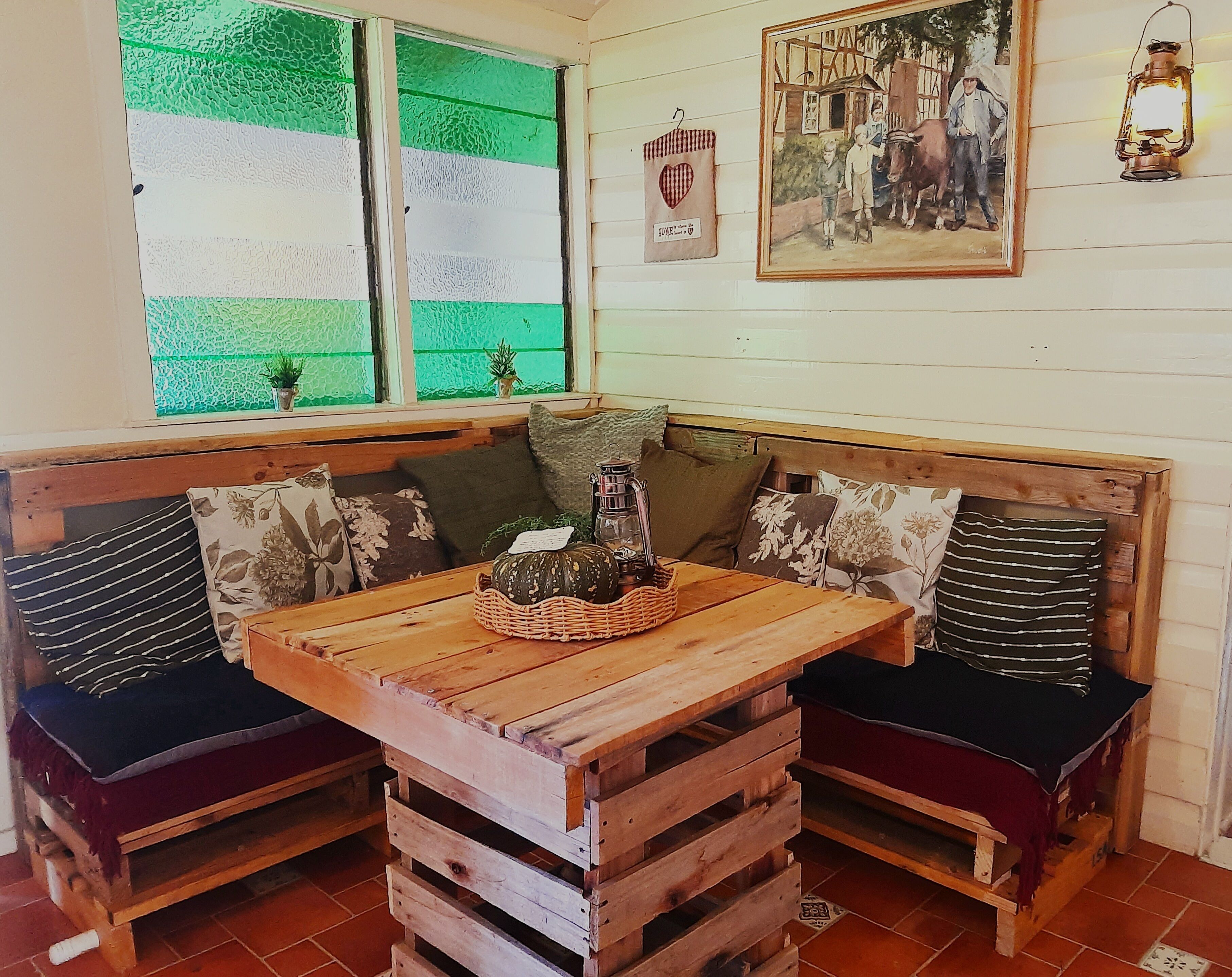 A rustic wooden bench seating area with a table in the center, a green window with a view, and a framed picture on the wall.