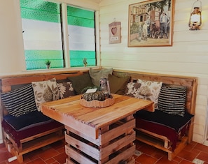 A rustic wooden bench seating area with a table in the center, a green window with a view, and a framed picture on the wall.
