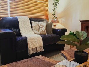 A living room with a dark leather sofa, a white throw blanket, and a patterned pillow. There is a floor lamp with a woven shade and a hanging plant with green leaves.