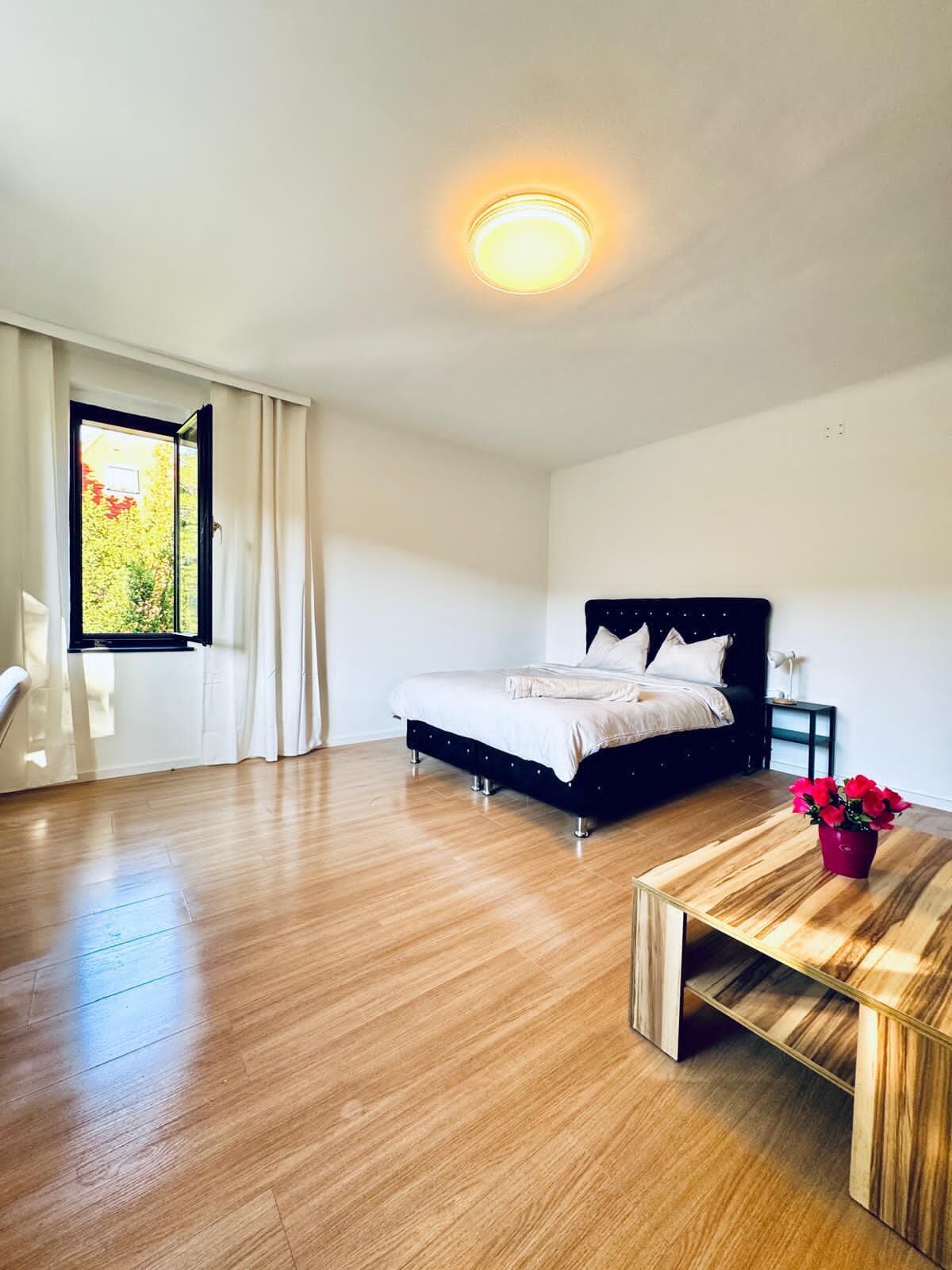 Un dormitorio con cama, una mesita de noche con un florero, una ventana con cortinas y una lámpara en el techo.