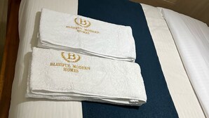 En pænt redt seng med hvide og marineblå sengetøj, to håndklæder med 'BLISSFUL MODERN HOMES' branding, og en hvid kurv på sengegavl.