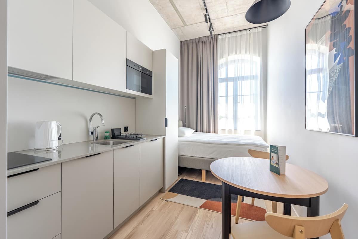 Une cuisine moderne dotée d’un four encastré, un coin repas avec une table et des chaises, un lit avec vue sur la fenêtre et un grand tableau abstrait accroché au mur.