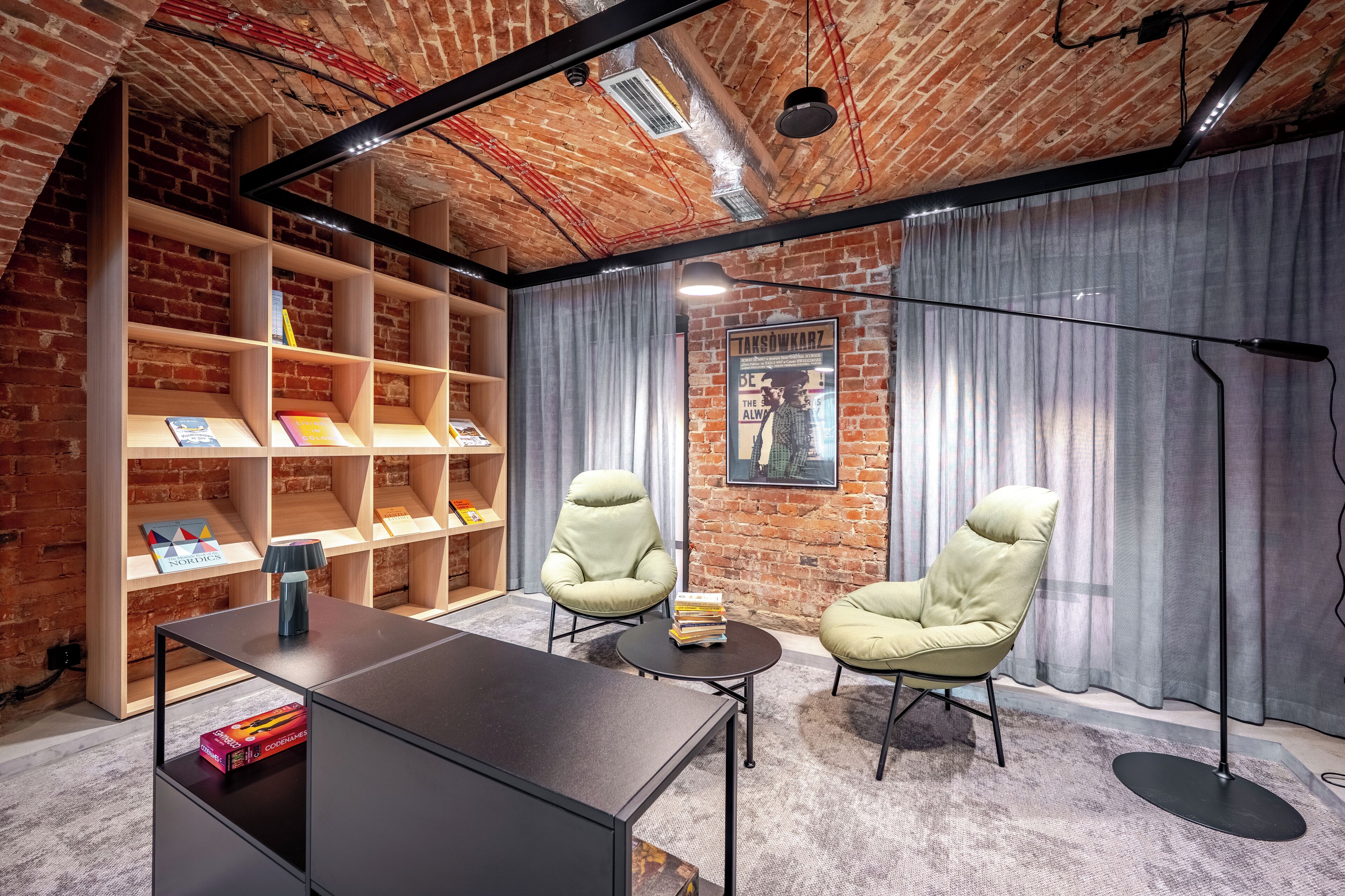 A room with exposed brick walls, a wooden bookshelf, two armchairs, a desk, and a floor lamp.