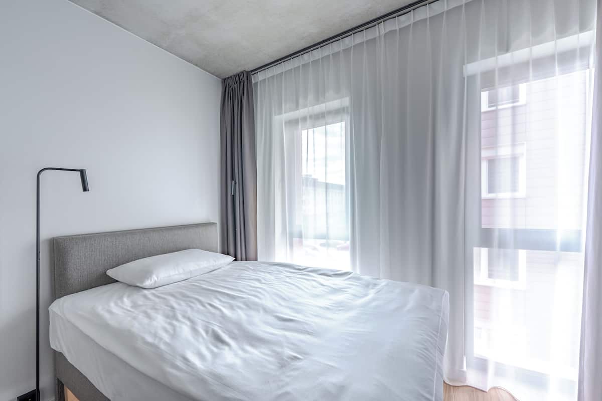 Une chambre d’hôtel moderne avec un grand lit, une table de chevet et une fenêtre ornée de rideaux légers.