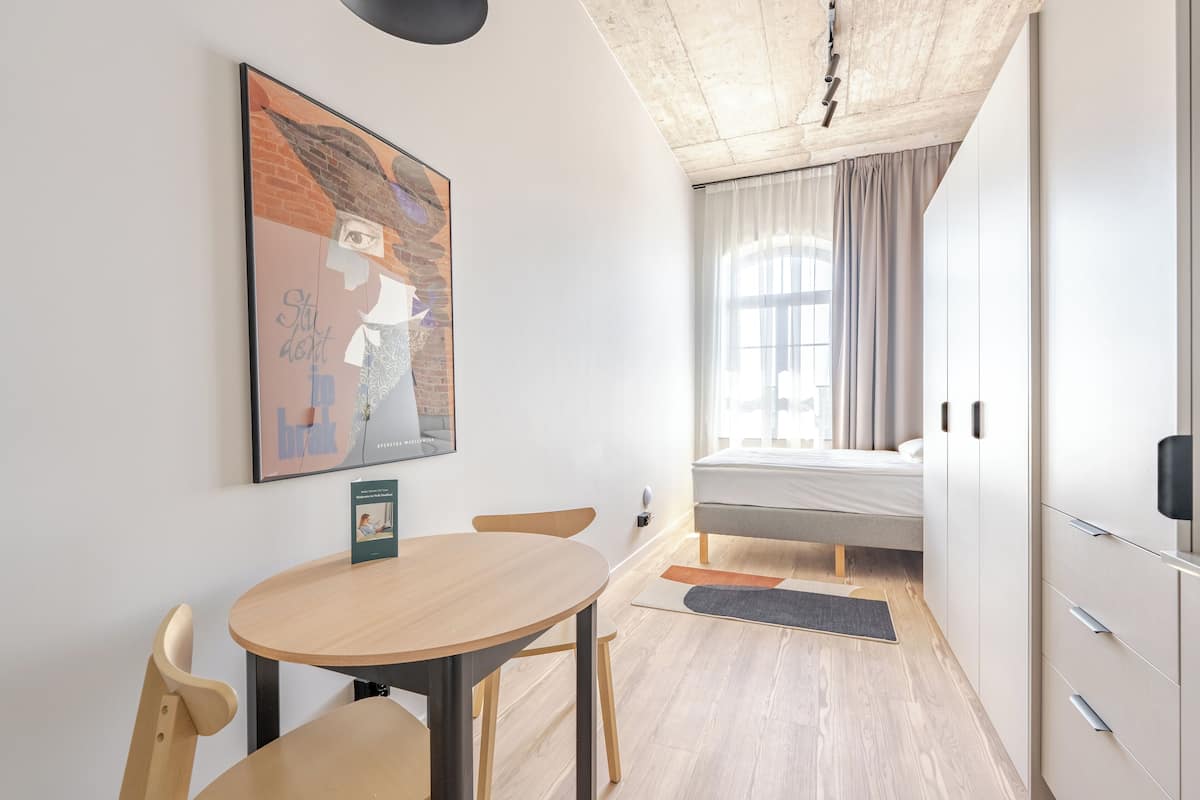 Une pièce de taille modeste, comprenant un lit, une petite table et une chaise. On y trouve une fenêtre avec des rideaux, une œuvre d’art accrochée au mur et une porte.