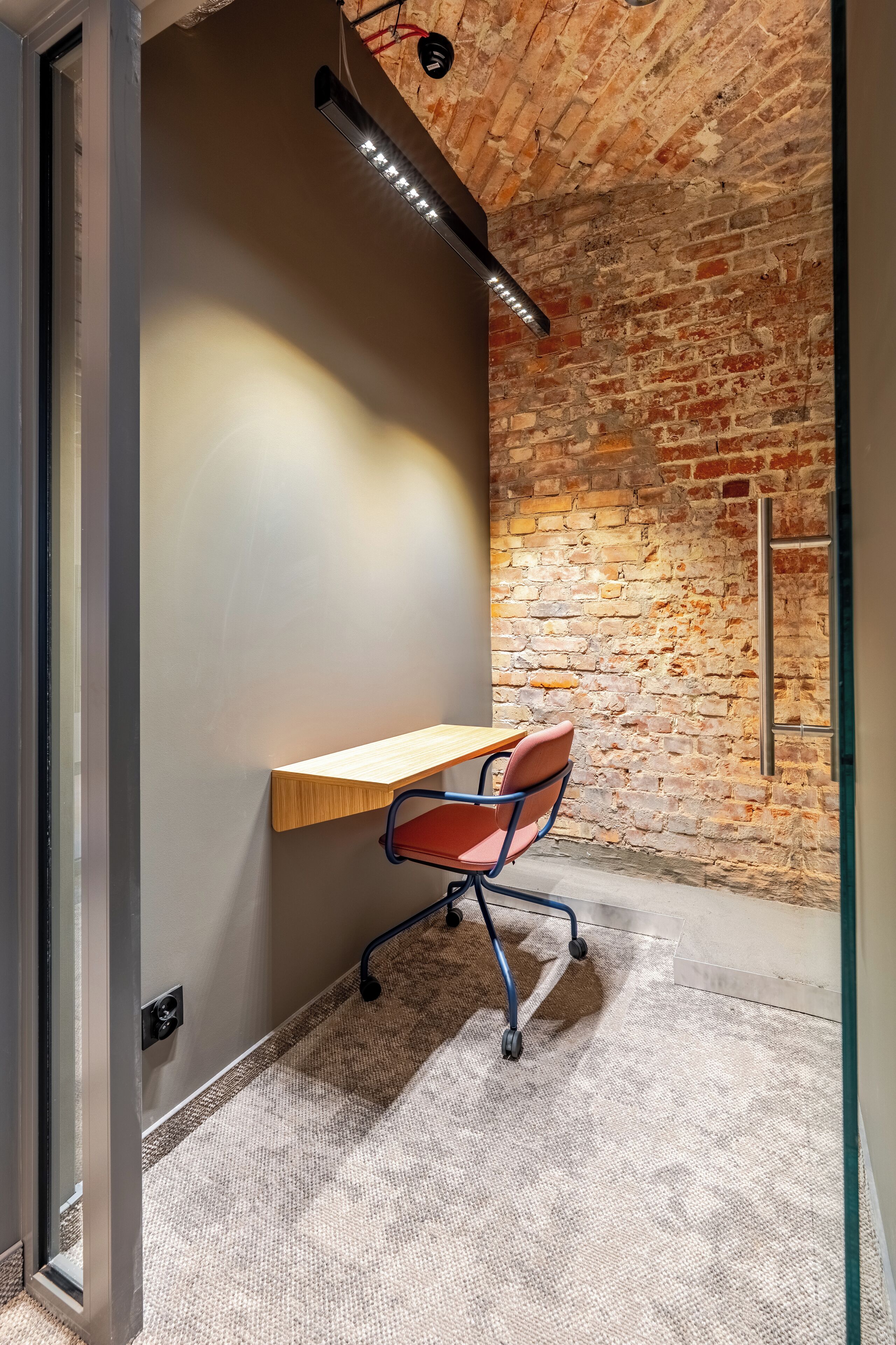 A room with a wooden desk, a red office chair, a brick wall, and a modern light fixture.