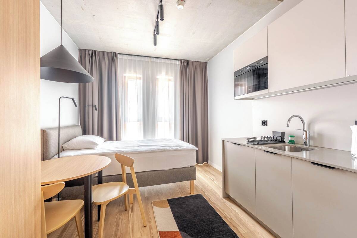 Une chambre d’hôtel de taille réduite, comprenant un lit, une petite table à manger, des chaises, un coin cuisine et une fenêtre avec des rideaux.
