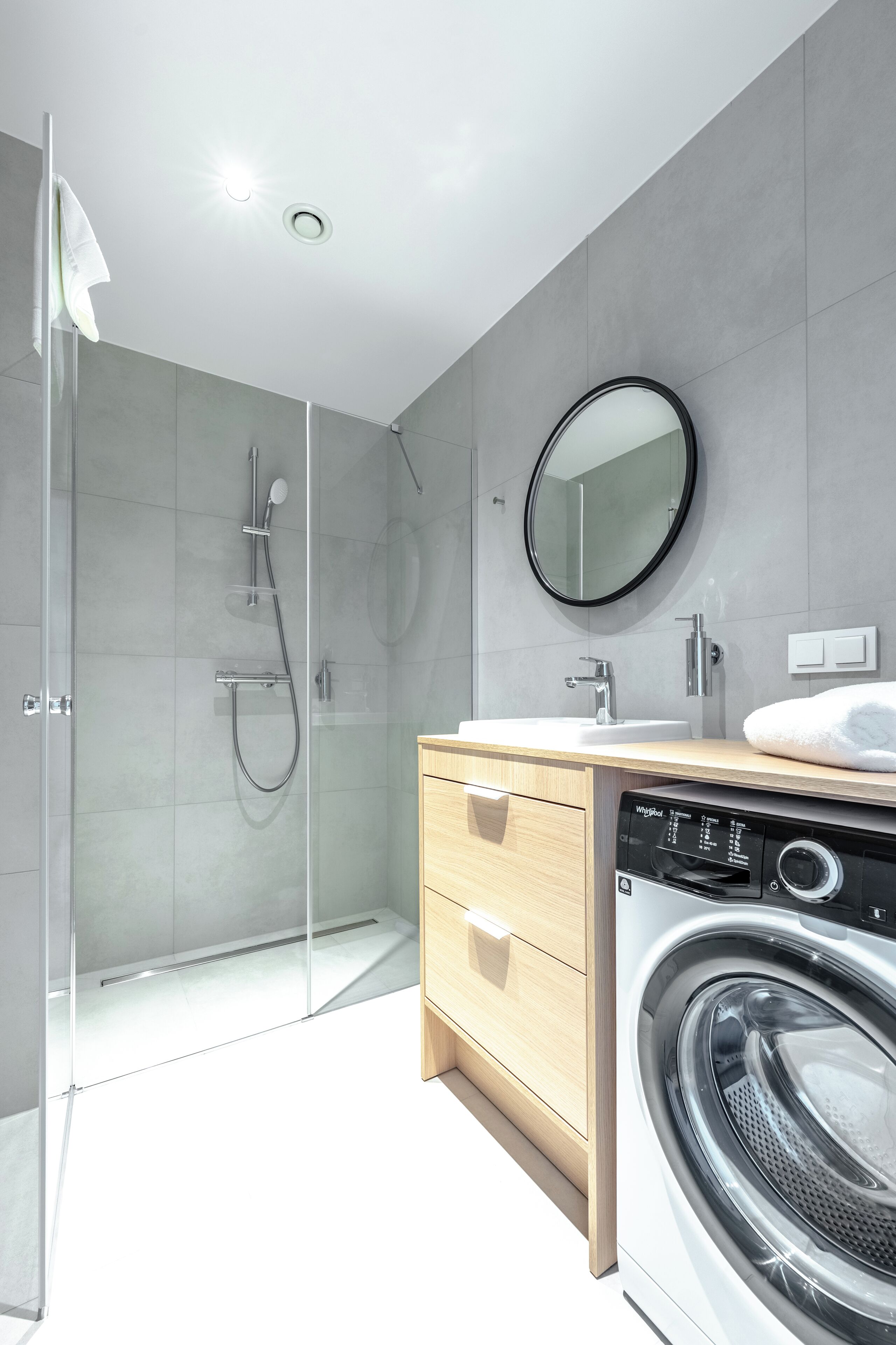 A modern bathroom with a glass-enclosed shower, a round mirror, a sink, and a built-in washing machine.