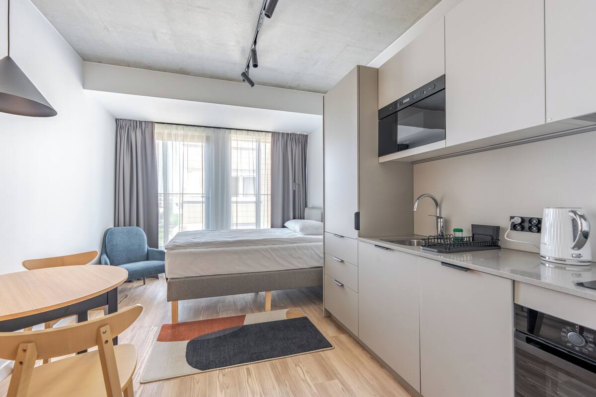 Une chambre d’hôtel moderne équipée d’un lit, d’un bureau, d’une chaise, d’un coin cuisine et d’une fenêtre avec des rideaux.