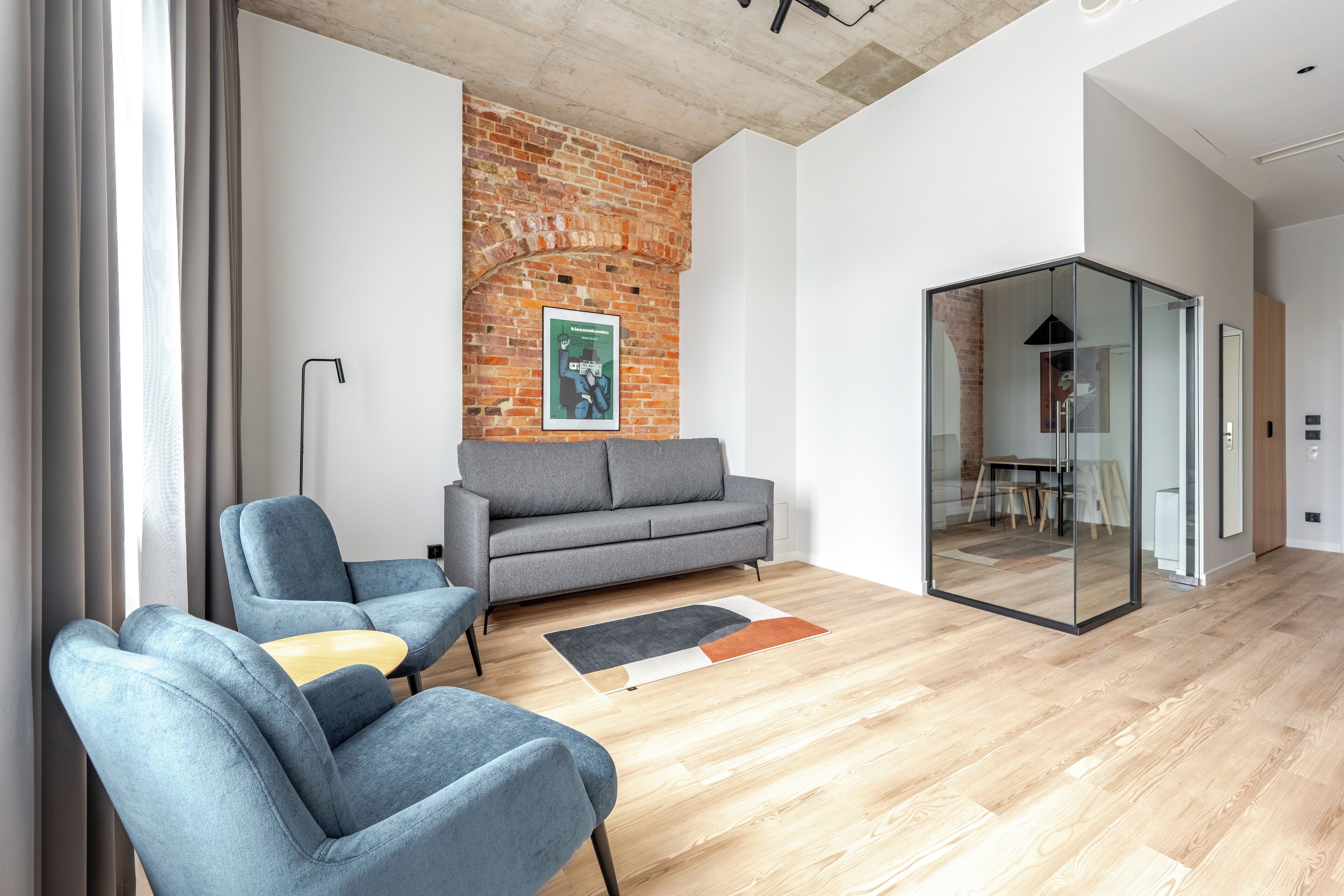 A modern living room with a grey sofa, blue armchairs, a wooden floor, and a brick wall.