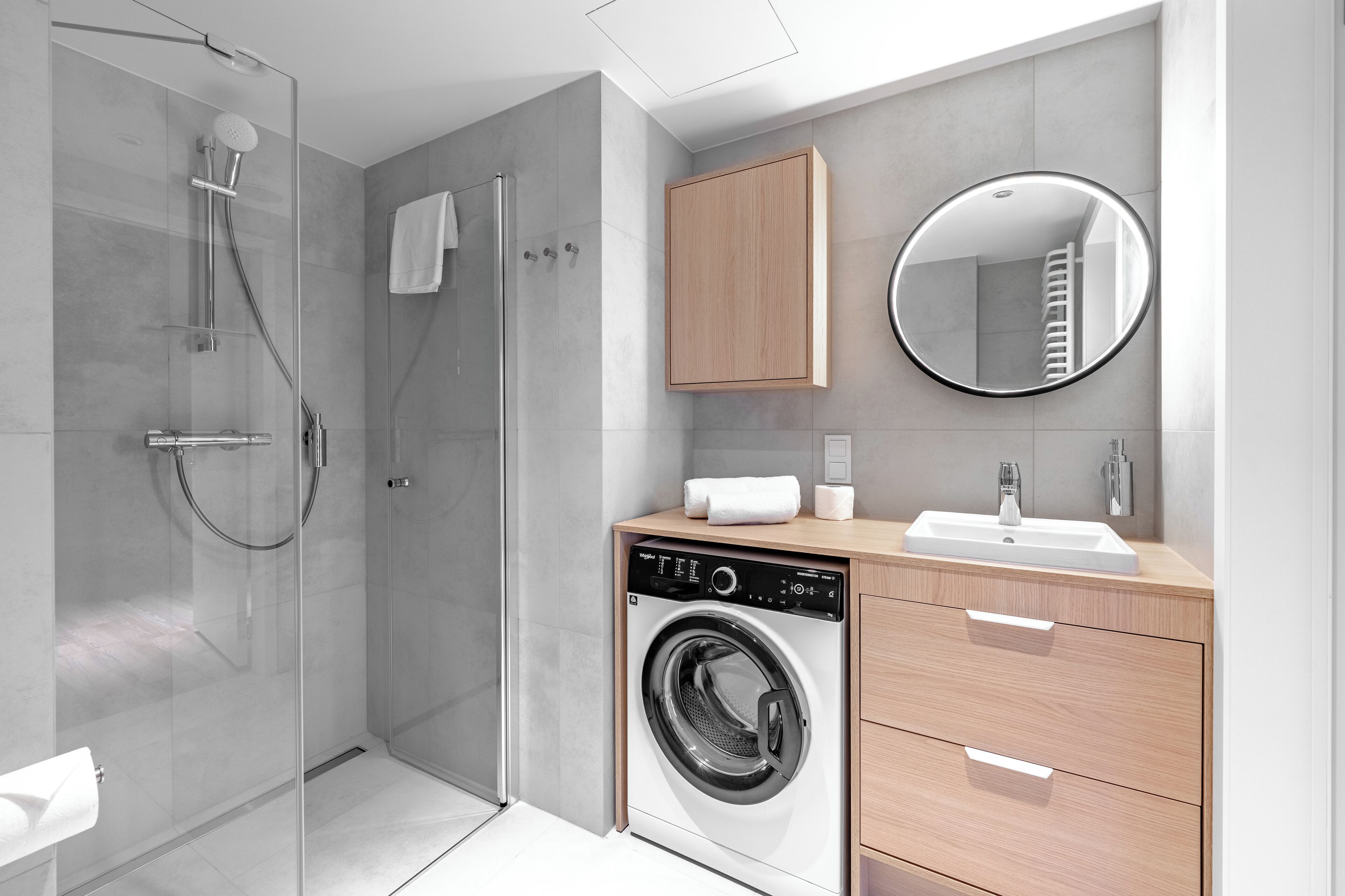 Un bagno moderno con doccia vetrata, lavatrice integrata, mobile bagno in legno con specchio e porta asciugamani.