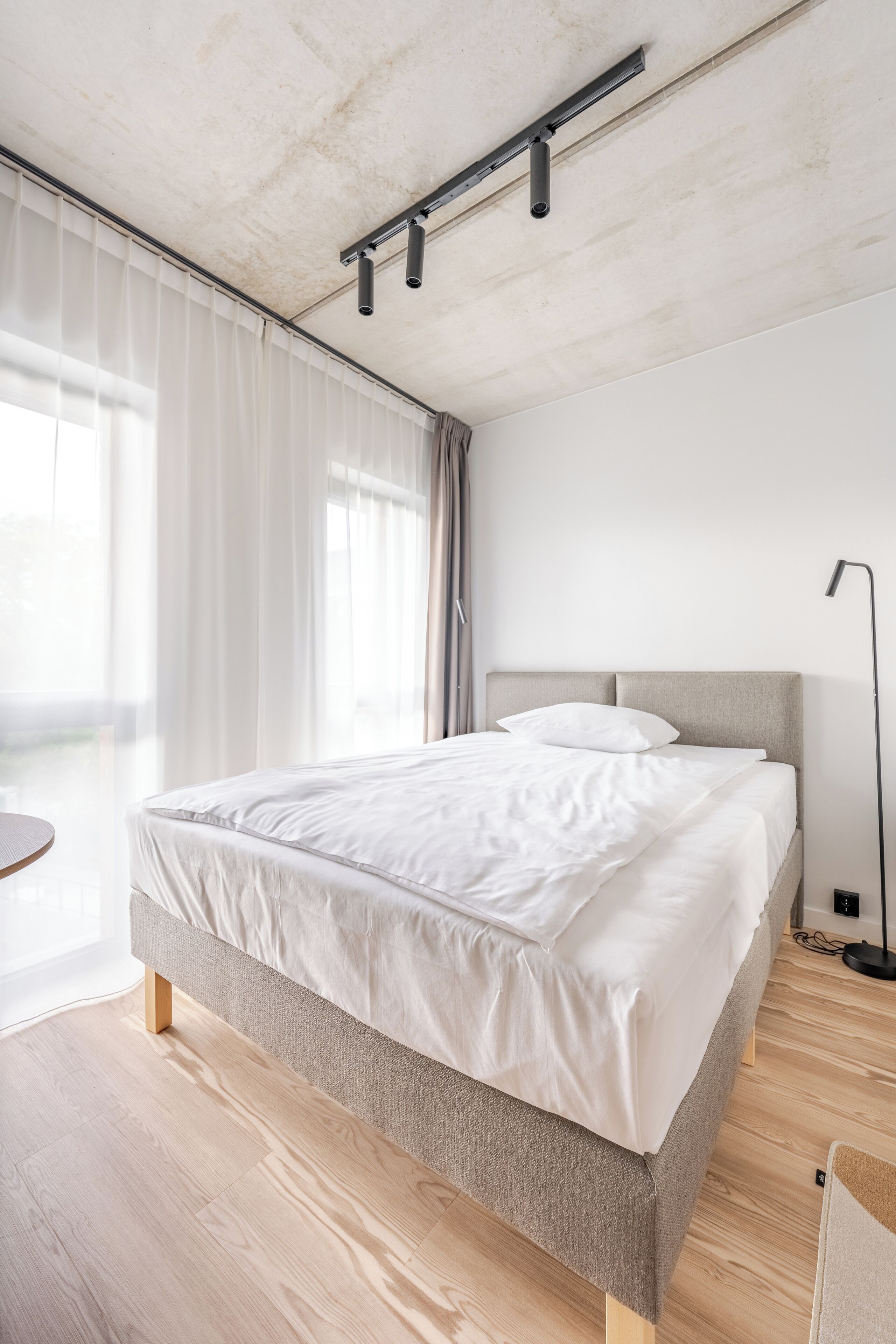 A modern bedroom with a large bed, a ceiling track lighting system, and a view of the outdoors through sheer curtains.