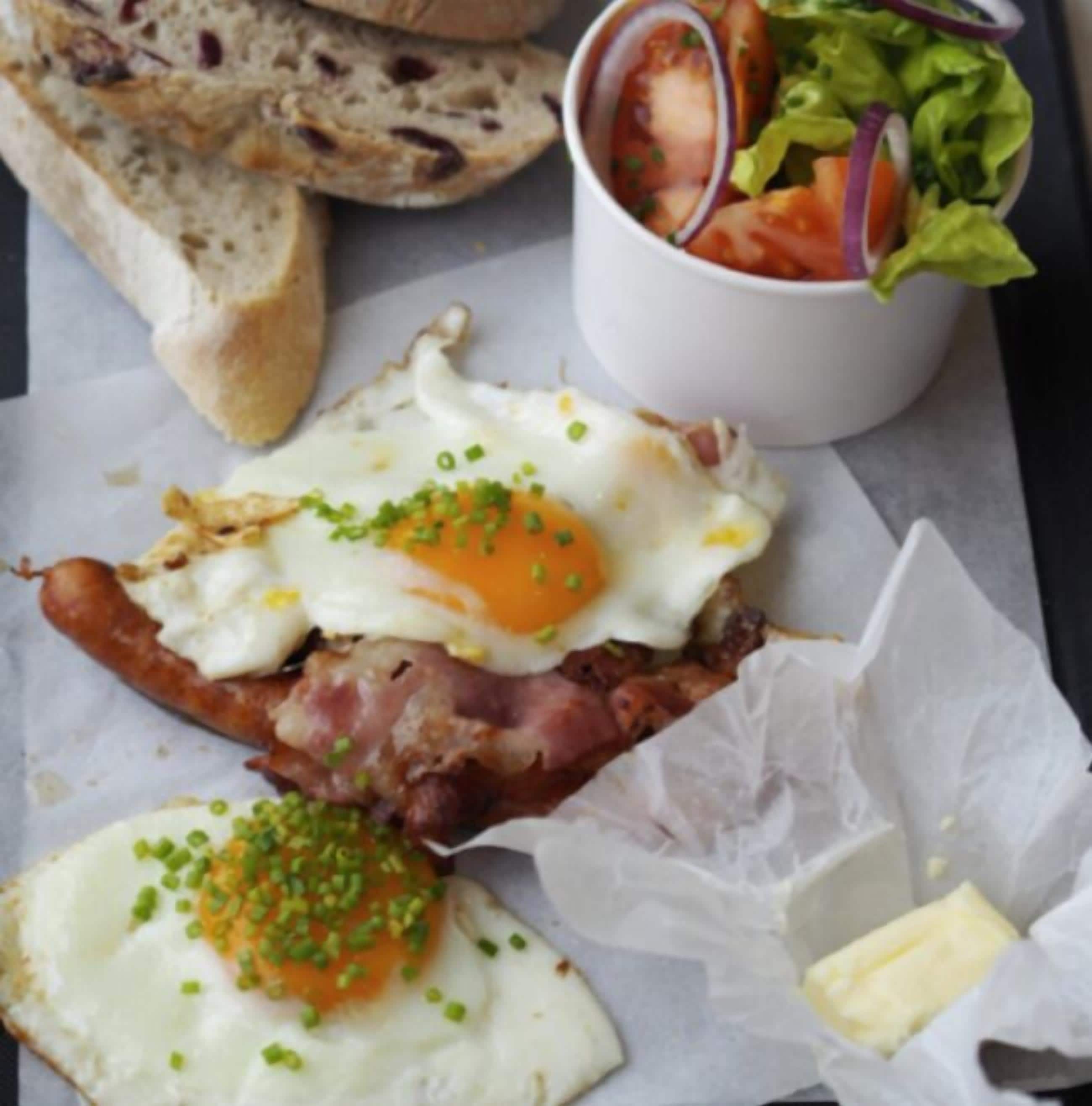 Un sándwich con huevo frito, tocino y cebollines sobre una rebanada de pan, acompañado de una ensalada pequeña y una taza de sopa de tomate.