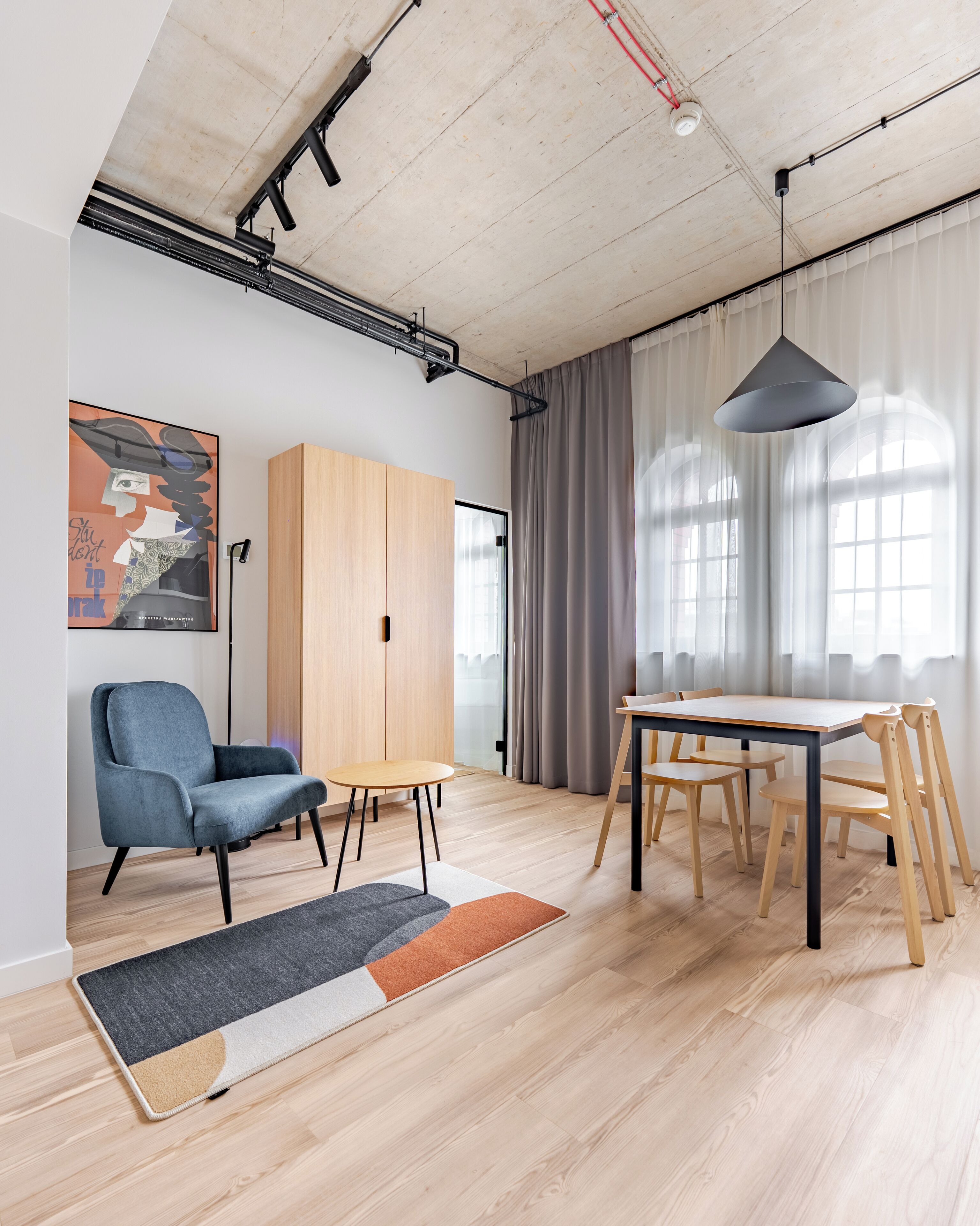 A modern interior with a wooden floor, a blue armchair, a wooden table with chairs, a hanging light fixture, and a large window with curtains.