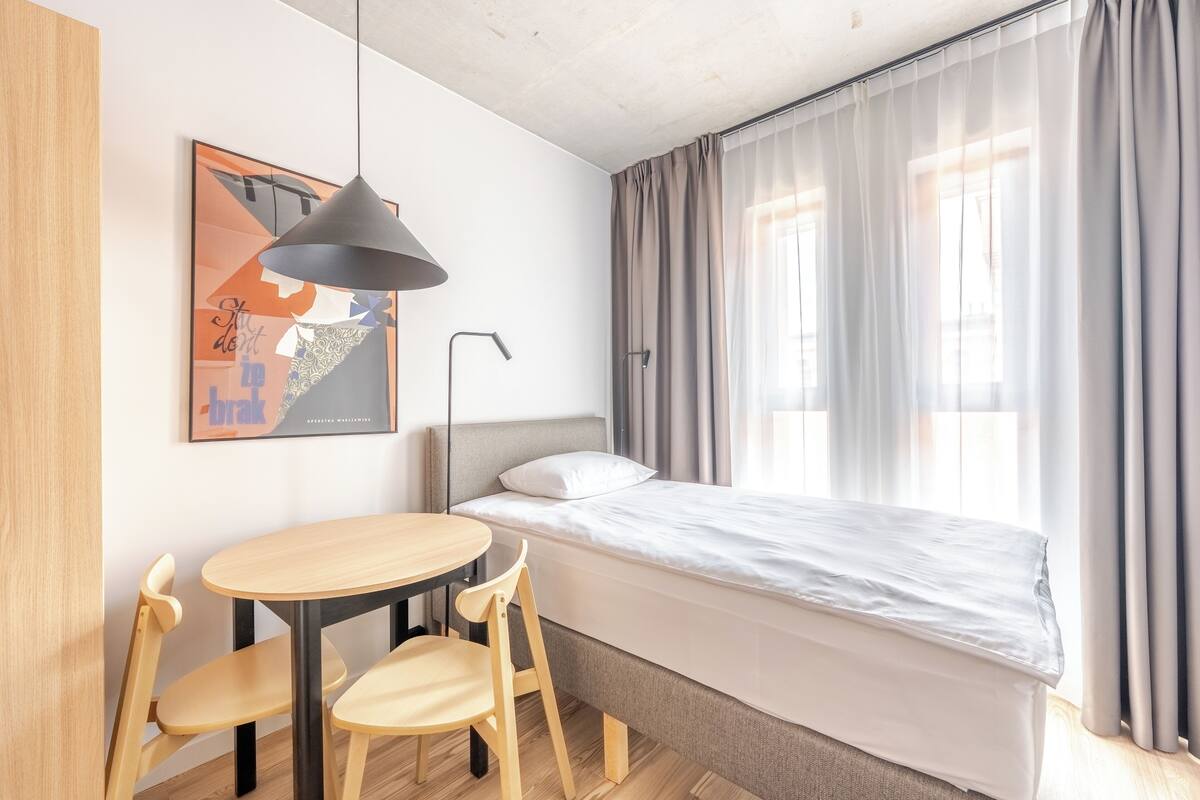 Une chambre à coucher comprenant un lit, un bureau, des chaises, une lampe suspendue et une fenêtre avec des rideaux.