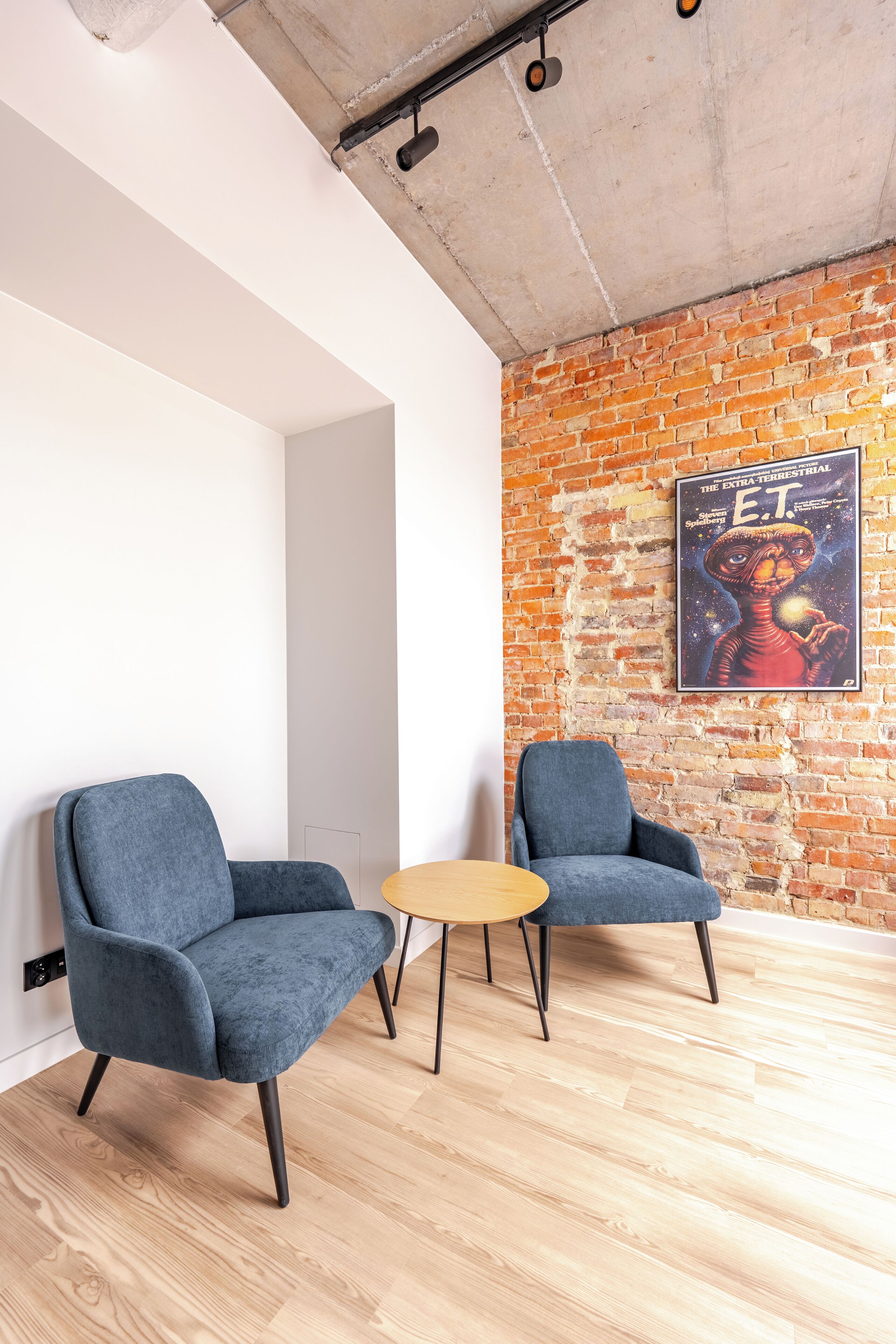 Ein Zimmer mit zwei grauen Sesseln, einem kleinen runden Tisch, einer Ziegelwand und einem Poster eines Science-Fiction-Charakters.