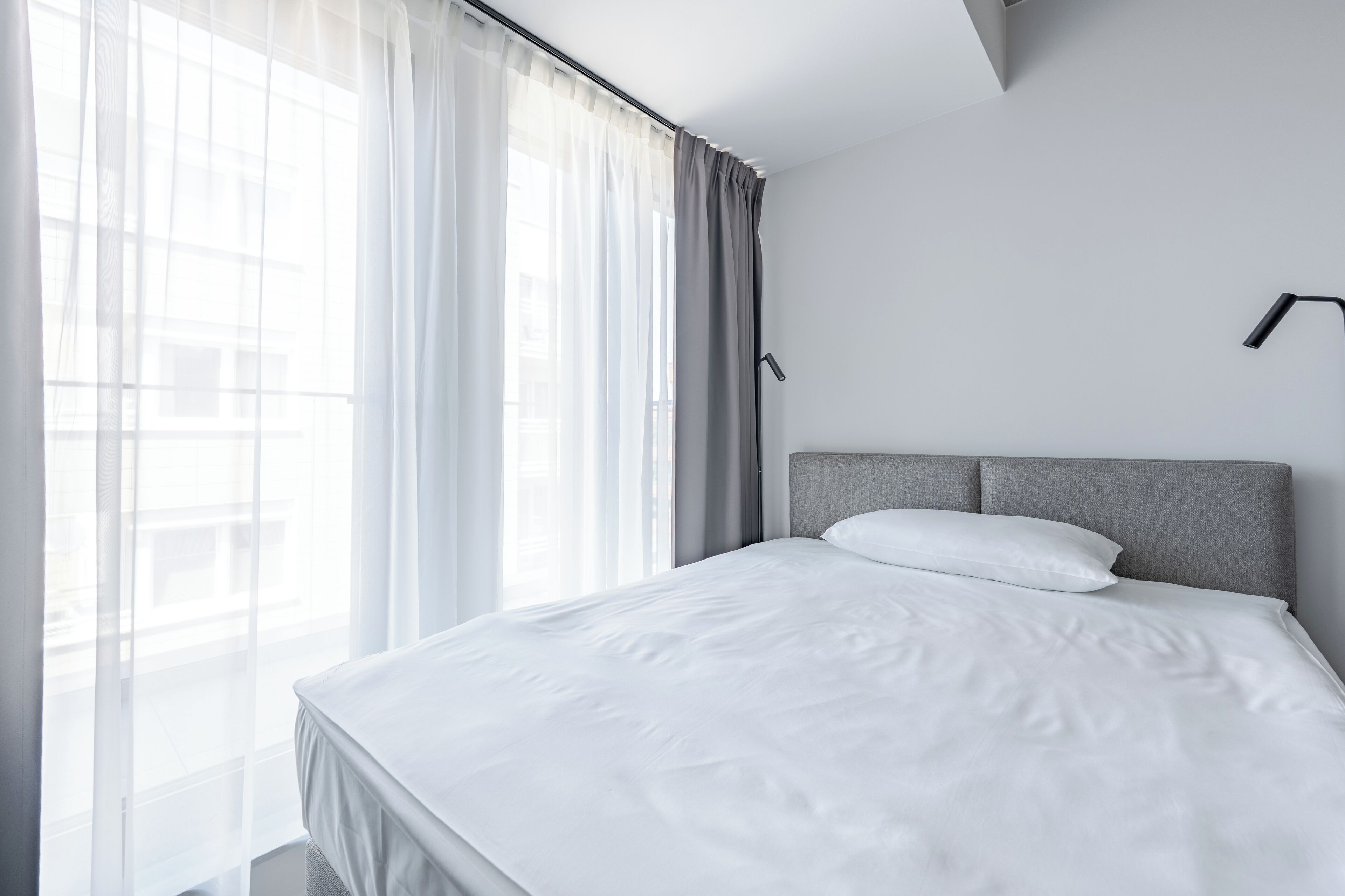 Una habitación de hotel moderna con una cama grande, un cabecero gris y cortinas translúcidas en una ventana.