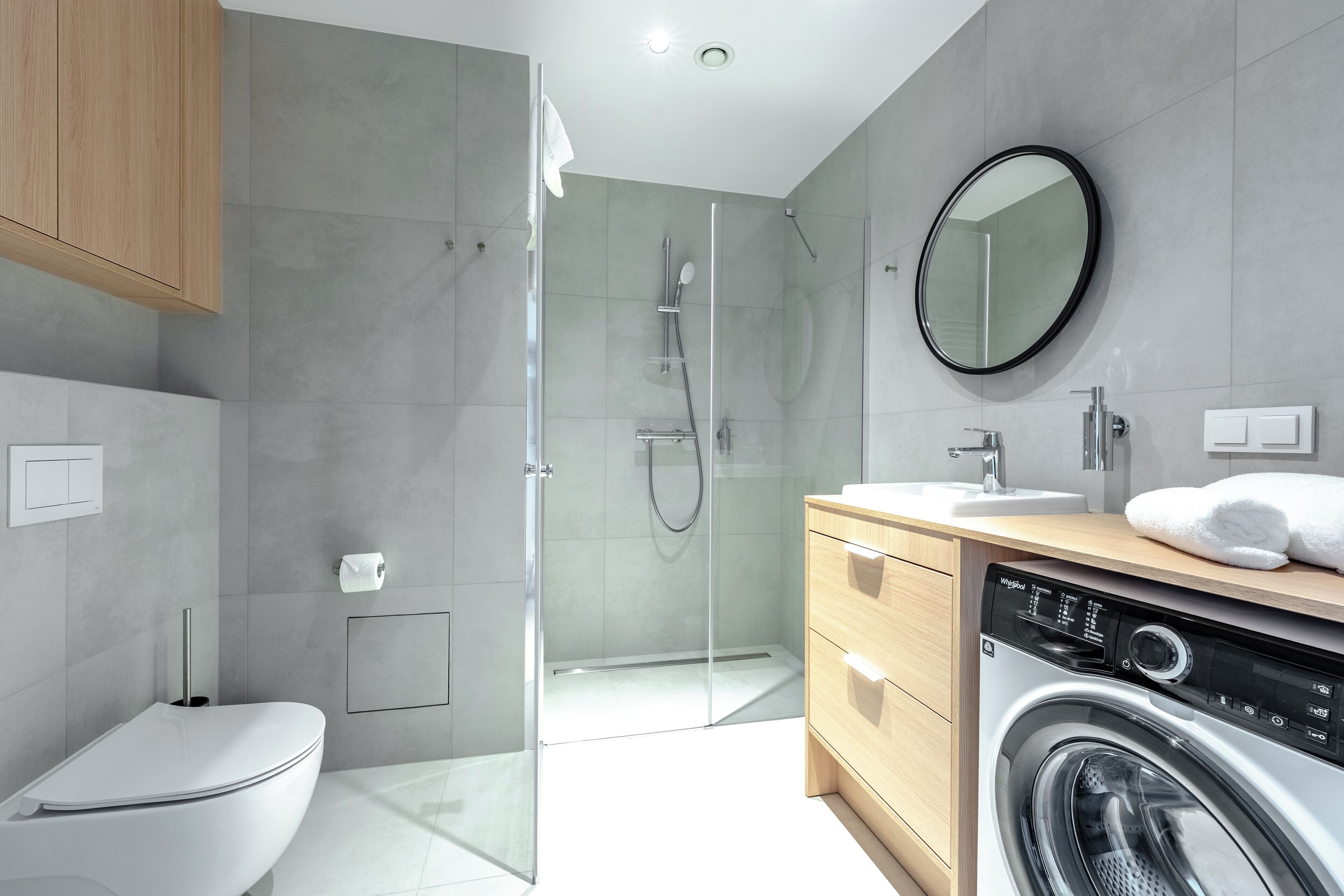 A modern bathroom with a glass-enclosed shower, a toilet, a washing machine, and a wooden vanity with a mirror.