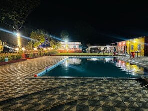 Une piscine dont le fond est recouvert de carrelage, entourée de bâtiments dont les fenêtres sont illuminées et dotés d’espaces de détente en plein air.