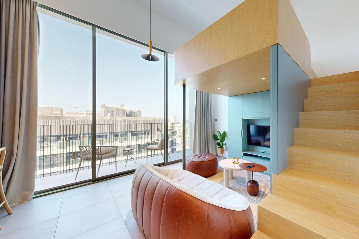 A modern living room with a large window, a brown leather sofa, a small round table, and a staircase.