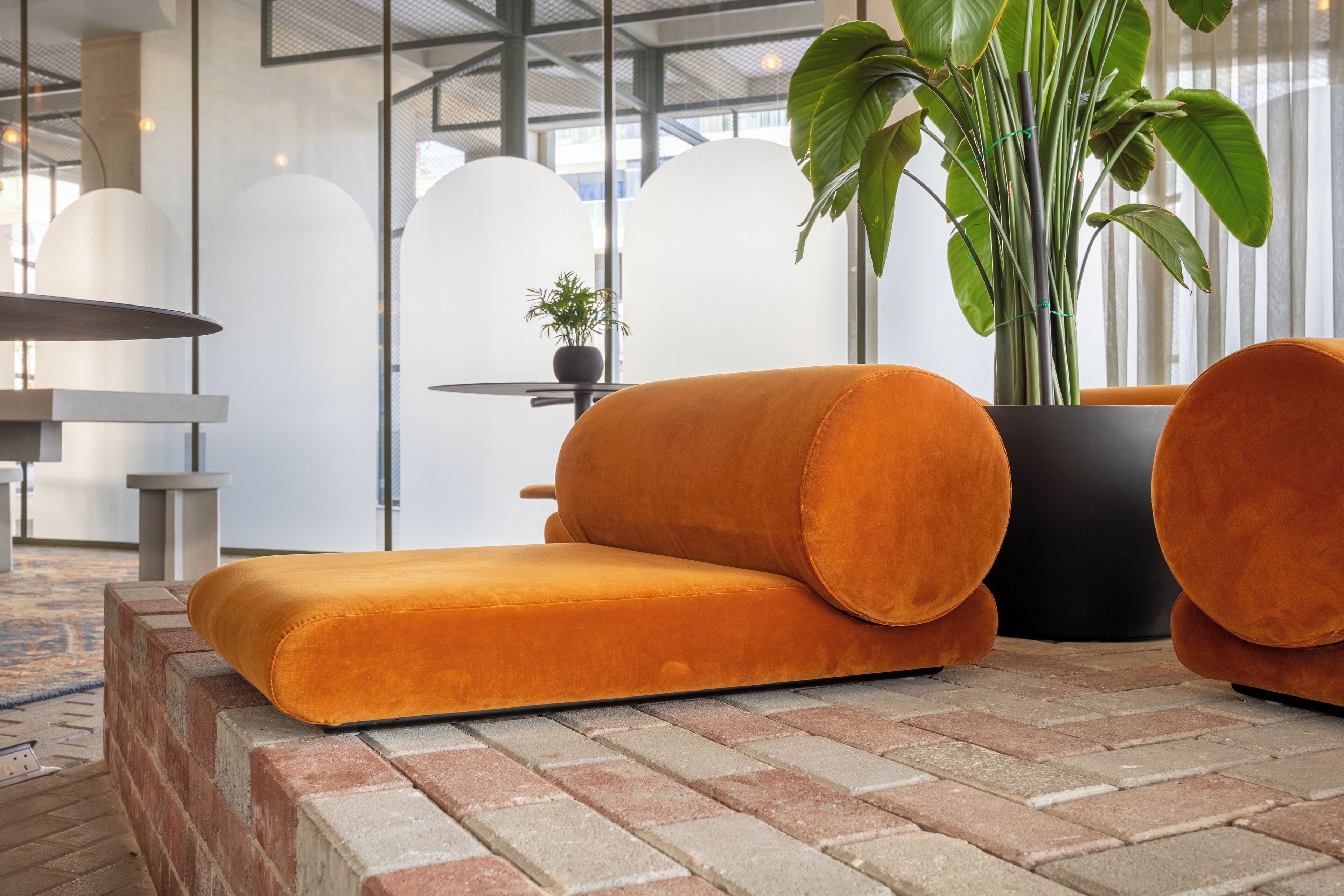 A modern interior with a brick floor, orange cushioned seating, a potted plant, and a glass partition.