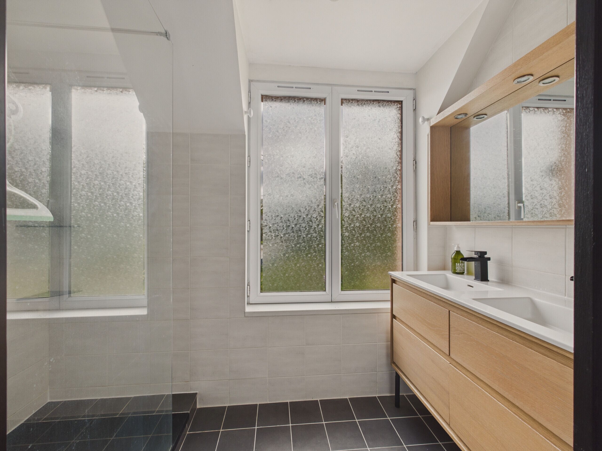 Een moderne badkamer met een douche, een raam met gezandstraald glas, een wastafel en een houten opbergmeubel.