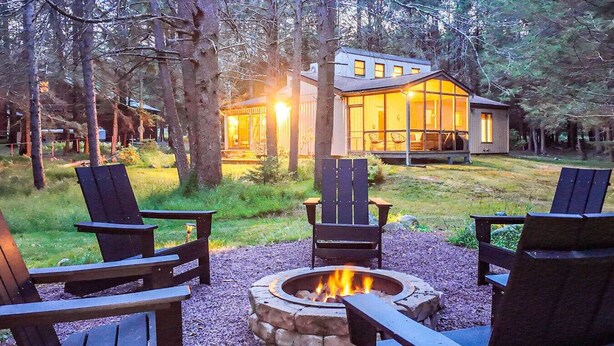 A cozy outdoor seating area with a fire pit, surrounded by Adirondack chairs, and a modern house in the background. - Tranquil  Pocono Escape (Blakeslee)