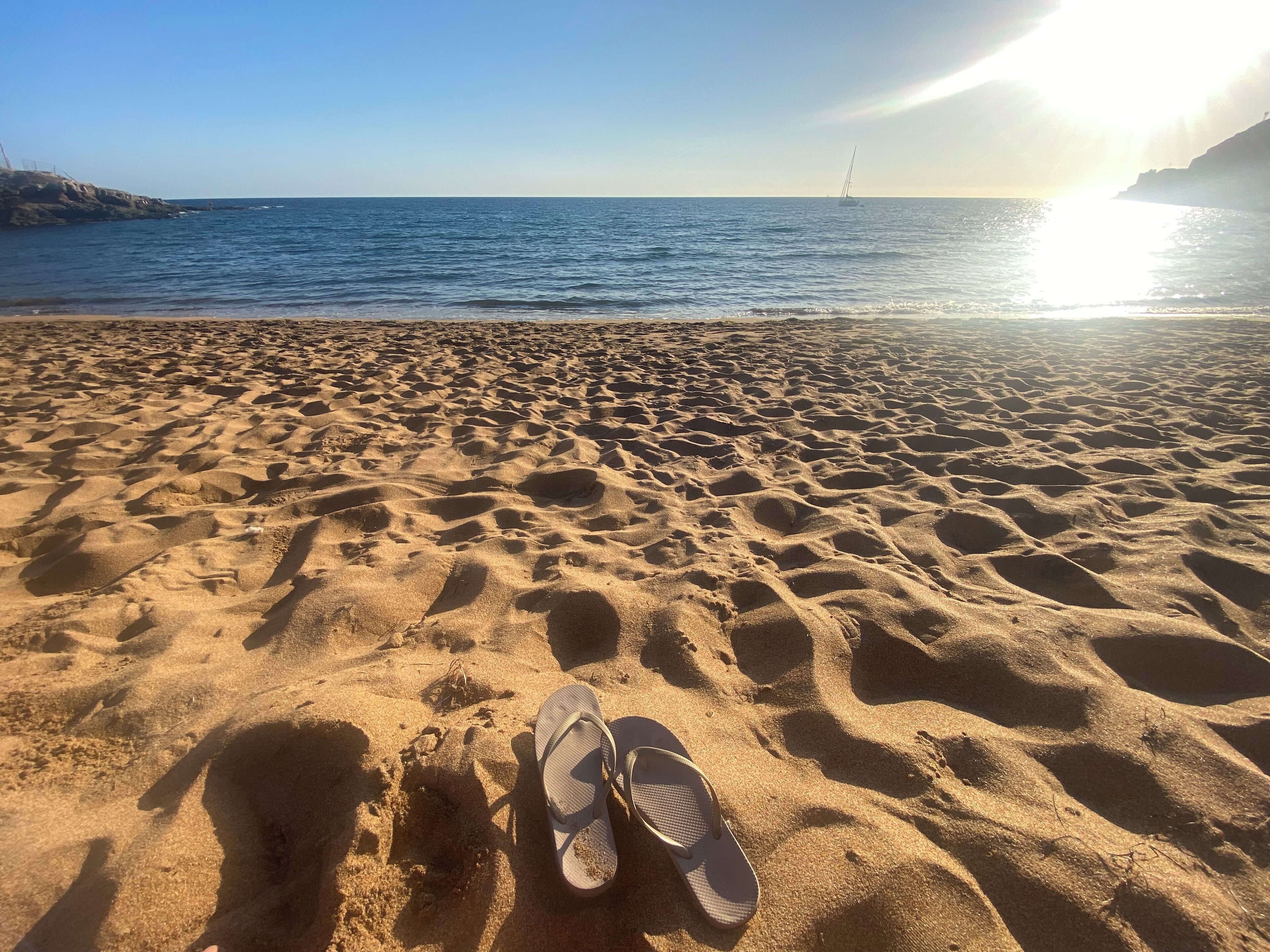 A beach with a pair of flip-flops on the sand and a sailboat in the water.