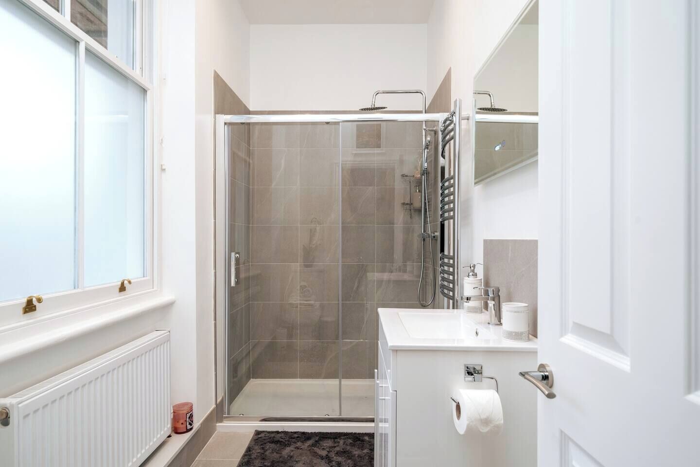 A modern bathroom with a glass shower enclosure, a white sink with a mirror, and a radiator.