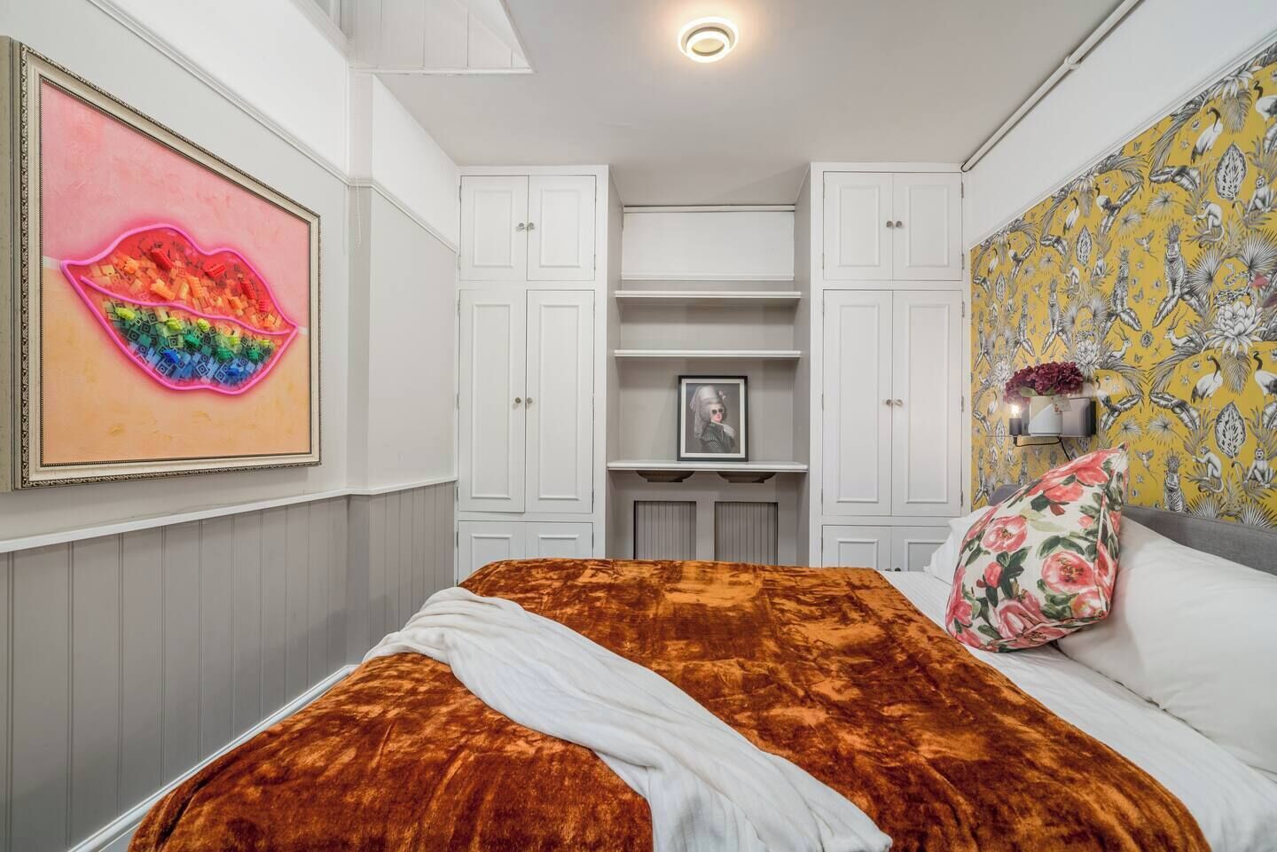 Ein Schlafzimmer mit einem Bett, einem Gemälde von Lippen, einem Blumenkissen und Tapete mit Muster.