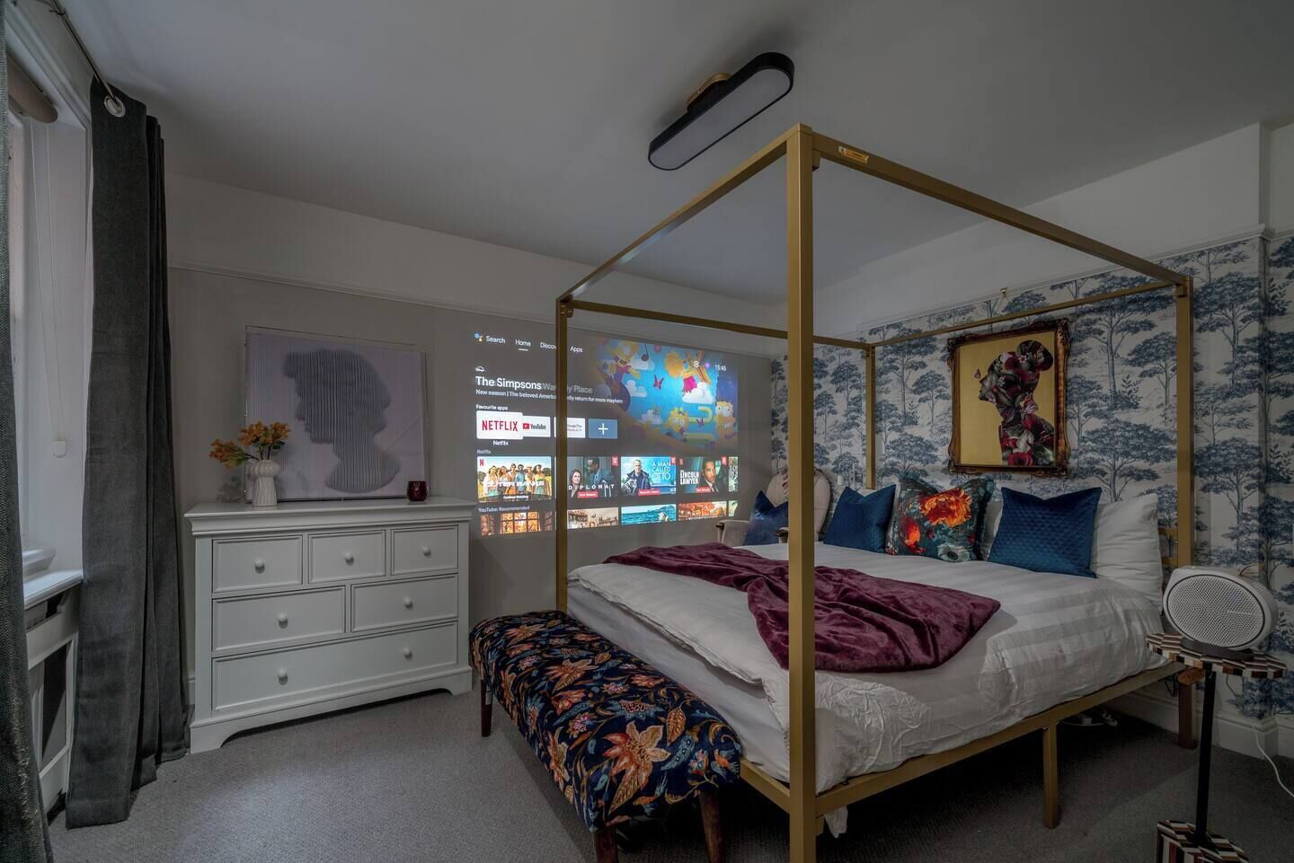 Ein Schlafzimmer mit Himmelbett, weißem Schminktisch, einer Bank und einem an der Wand befestigten Fernseher, der verschiedene Sender zeigt.