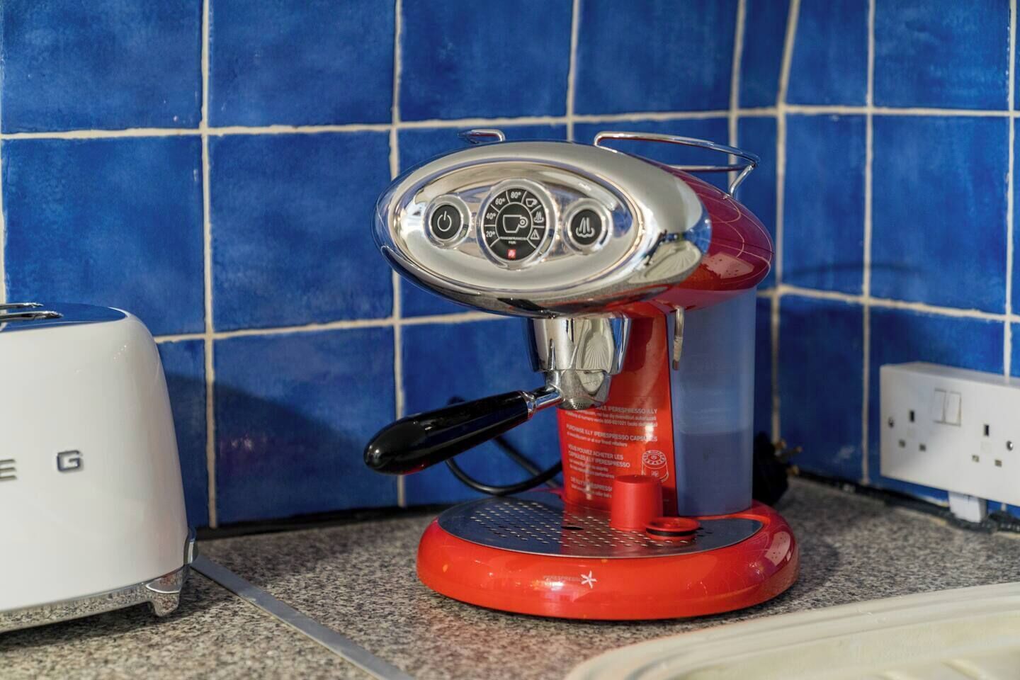 Een rode espressomachine met een digitaal display en een zwart handvat, geplaatst op een keukentrapje met blauwe tegels op de achtergrond.