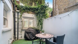 A small outdoor patio with a round table and two chairs, surrounded by a white wall and a brick building.